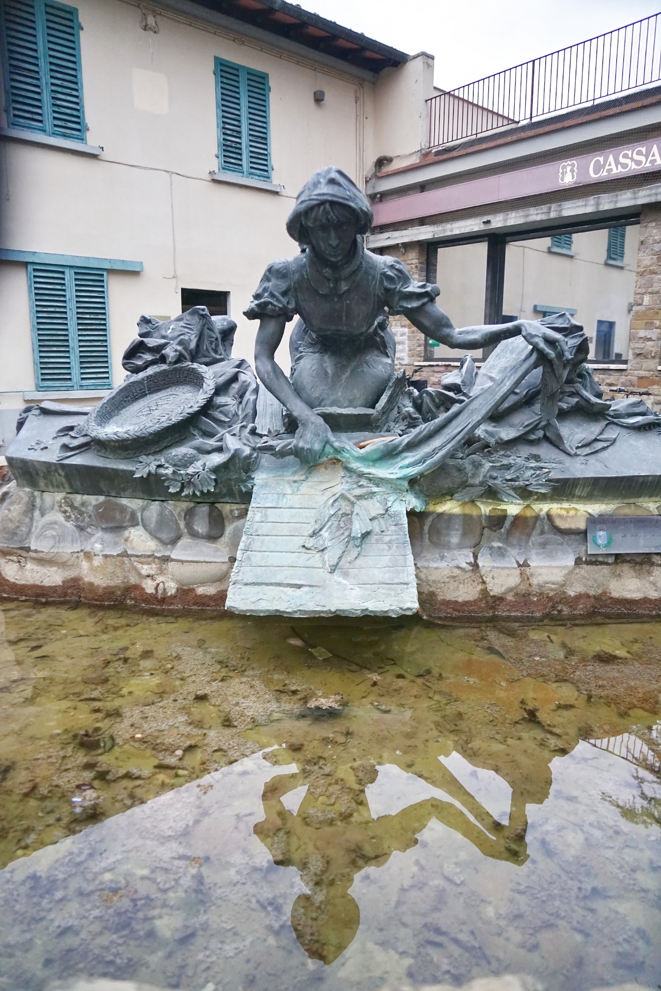 Statua in bronzo del monumento alla Lavandaia a Grassina, raffigurante una donna inginocchiata intenta a lavare i panni su un'asse di legno sopra una vasca d'acqua.