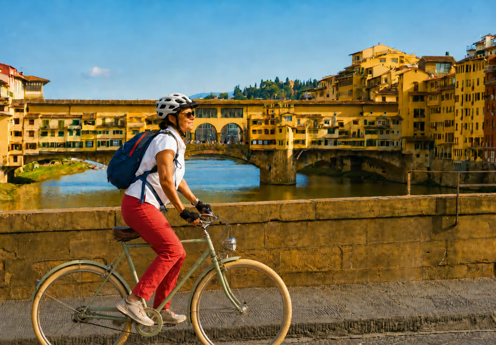 Woman on a vintage sage-green bicycle with helmet and backpack pedals along the Arno River in Florence, with the Ponte Vecchio and its characteristic yellow shops overlooking the Arno in the background.