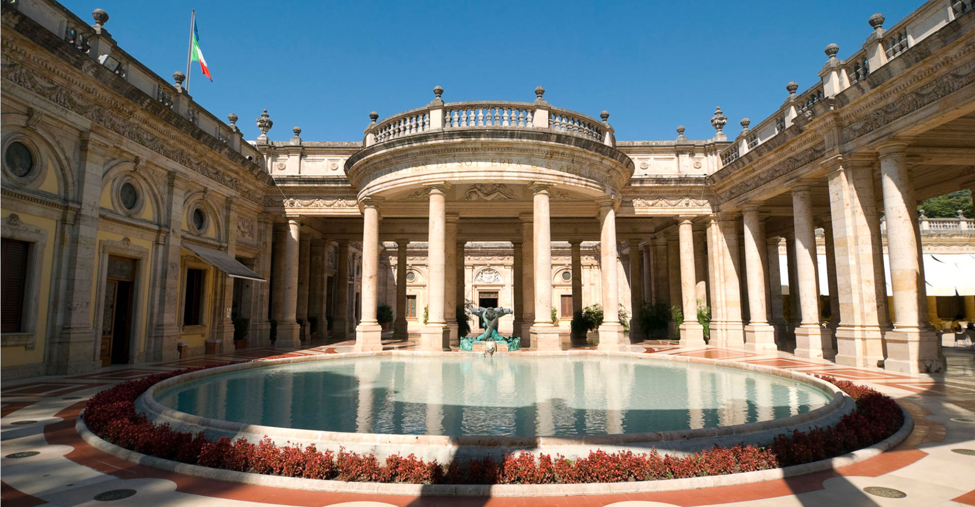The monumental courtyard of the Terme Tettuccio in Montecatini Terme with its central circular pool, bronze fountain, neoclassical colonnade, and Italian flag, home of the Montecatini Gender Reveal Experience