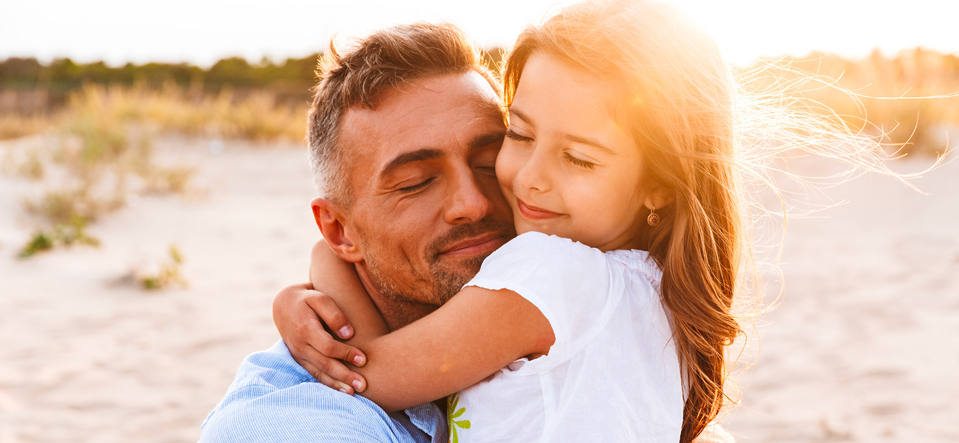 Papà single abbraccia sorridendo la figlia bambina su una spiaggia in Toscana al tramonto, momento di tenerezza durante un weekend in family hotel a Montecatini Terme