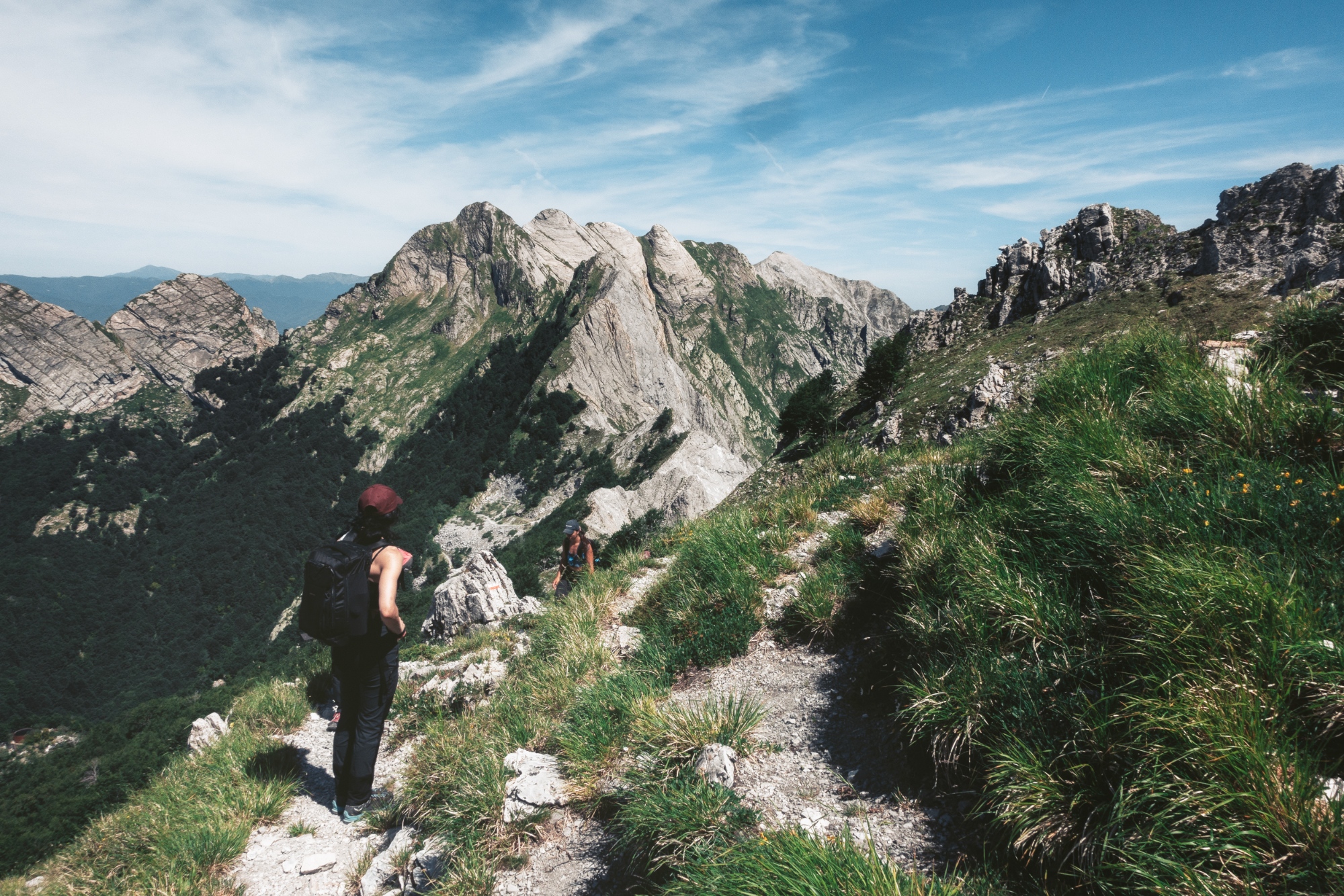 scursionisti con zaino in spalla percorrono un sentiero di cresta erboso sulle Alpi Apuane, con le imponenti pareti rocciose del Monte Cavallo e del Monte Contrario sullo sfondo e un cielo azzurro con nuvole sottili
