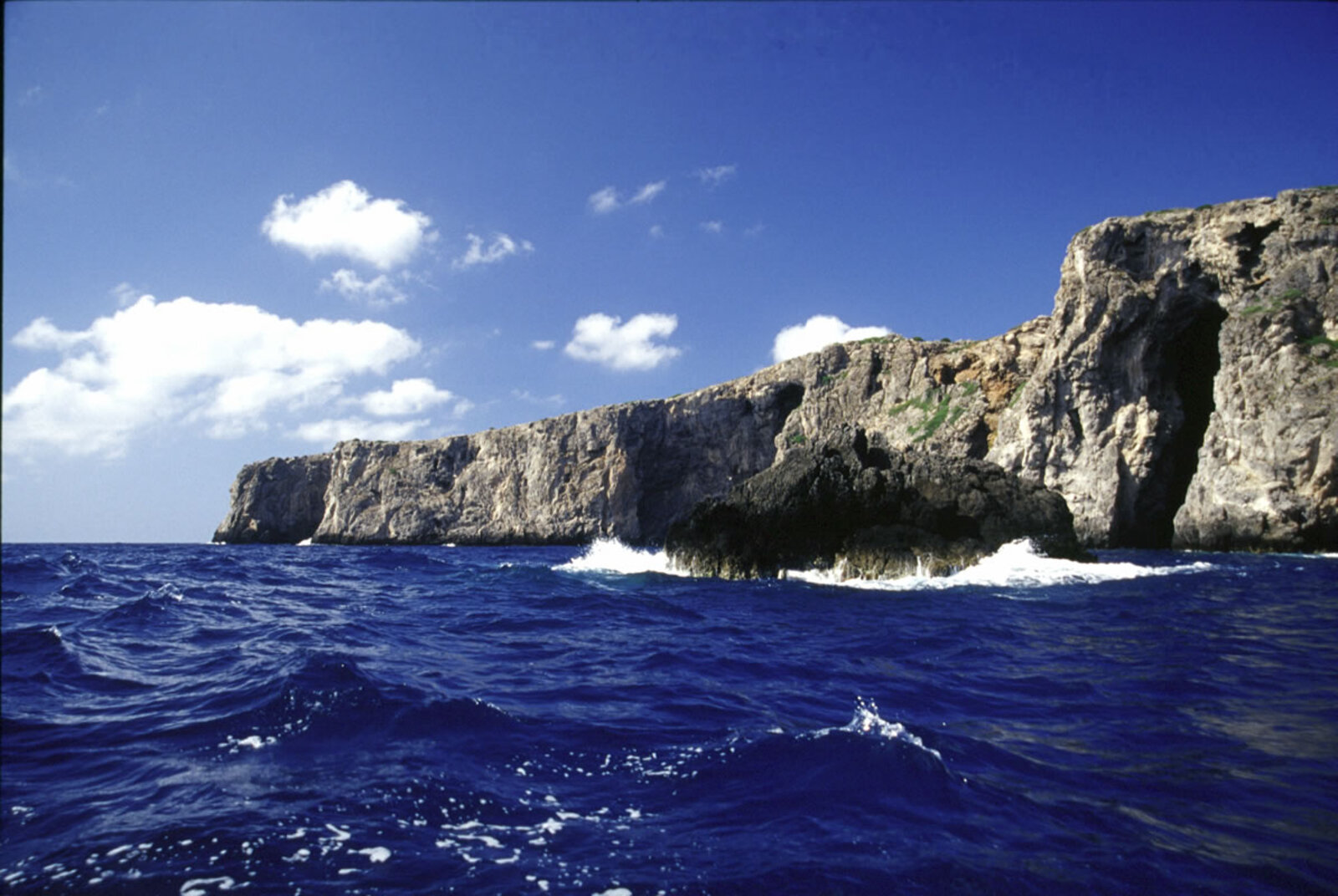 Falesie rocciose a picco sul mare all'Isola di Giannutri, con onde che si infrangono tra gli scogli vicino ai Grottoni di Punta Capel Rosso, cielo azzurro e nuvole sullo sfondo