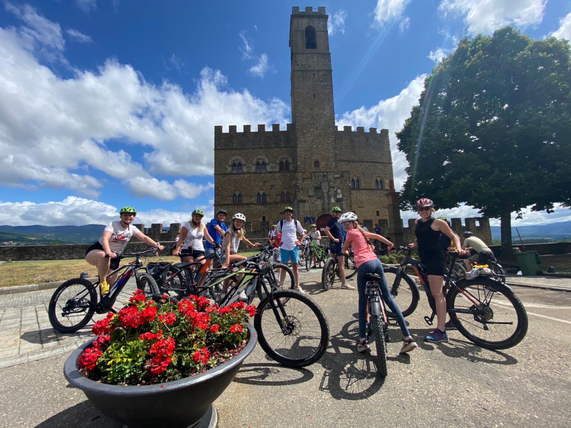 Group of cyclists with e-bikes and helmets in front of Poppi Castle, with a crenellated medieval tower in the background and a flowerbed with red geraniums in the foreground, during the tour from Poppi to Croce del Pratomagno.