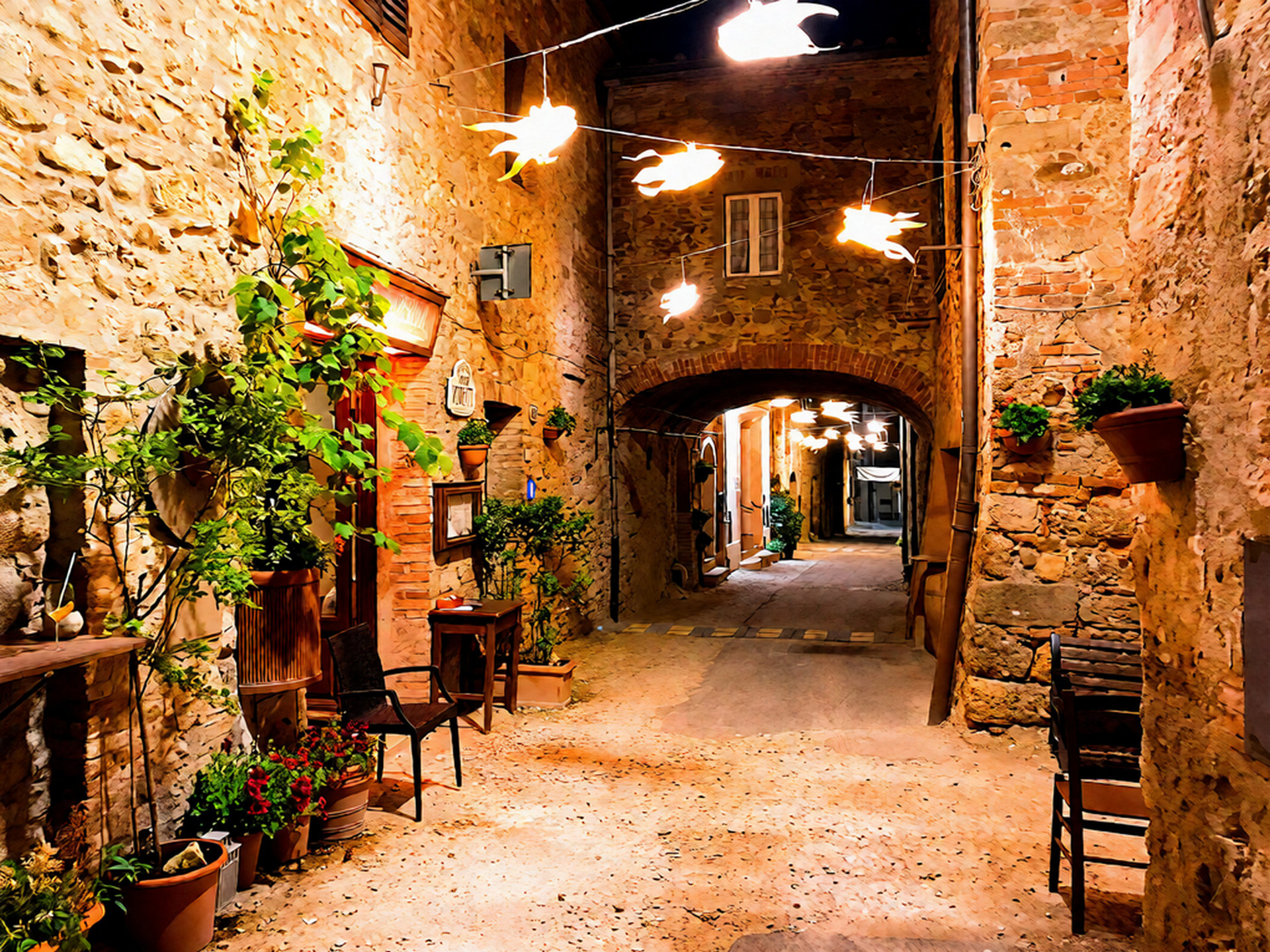 Vicolo medievale di Radicondoli illuminato di sera con luci decorative sospese, muri in pietra, archi e vasi di piante lungo il passaggio