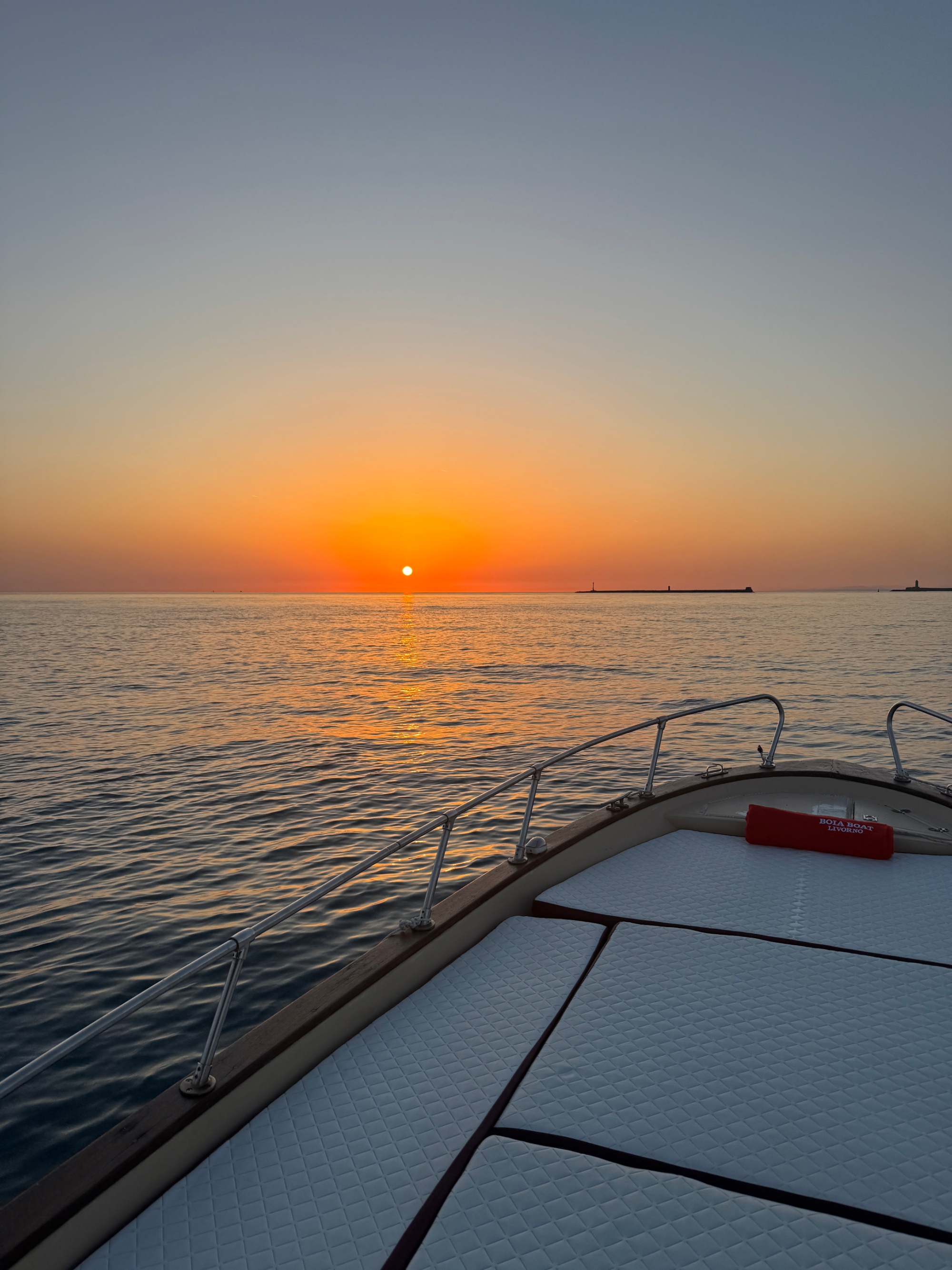 Prua della barca Boia Boat Livorno con asciugamano rosso brandizzato sul prendisole bianco, ripresa durante un tramonto sul mare con cielo arancione e sagoma del porto di Livorno all'orizzonte