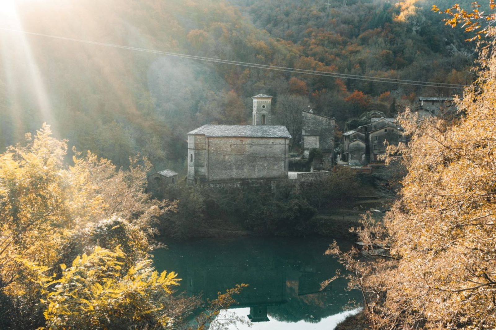 Chiesa in pietra con campanile circondata da boschi autunnali color oro che si riflettono in un lago turchese di montagna
