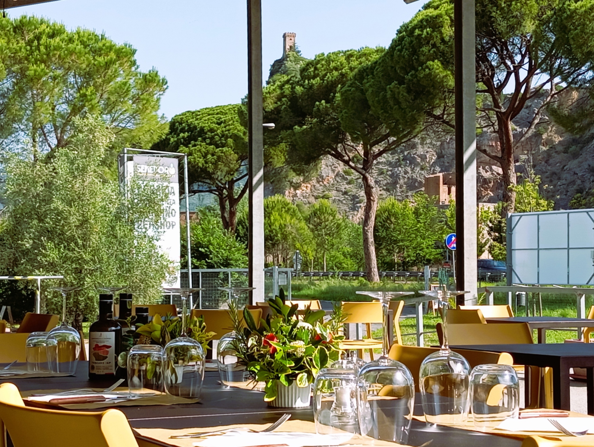 Tables set with glasses and floral centerpieces on the covered terrace of the Ristoro del Frantoio del Monte Pisano, overlooking maritime pines and the medieval tower of Vicopisano