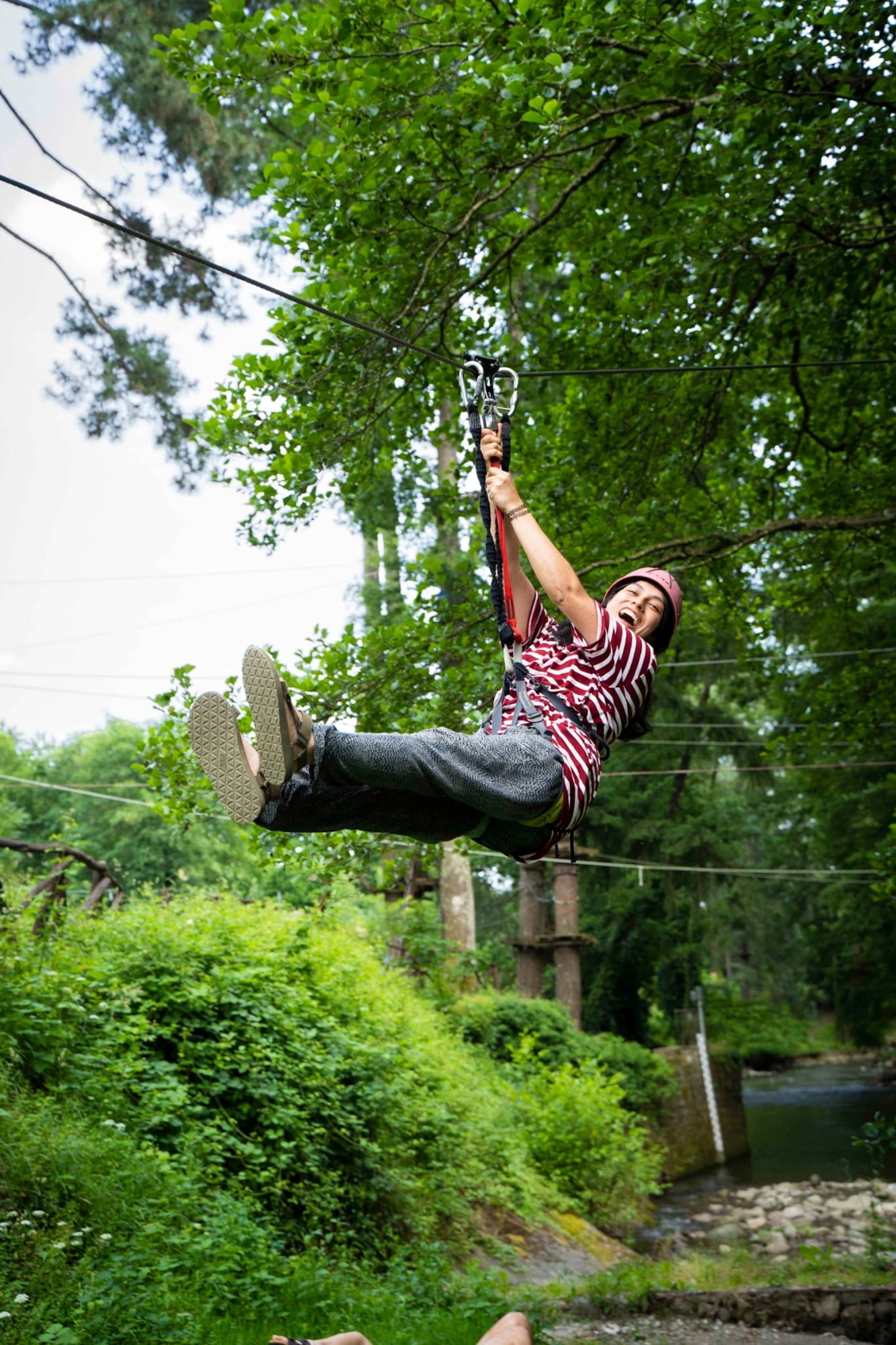 Smiling woman wearing a pink helmet and harness waving as she slides on a zipline over the river, immersed in the woods of the Canto alla Rana river park in Stia