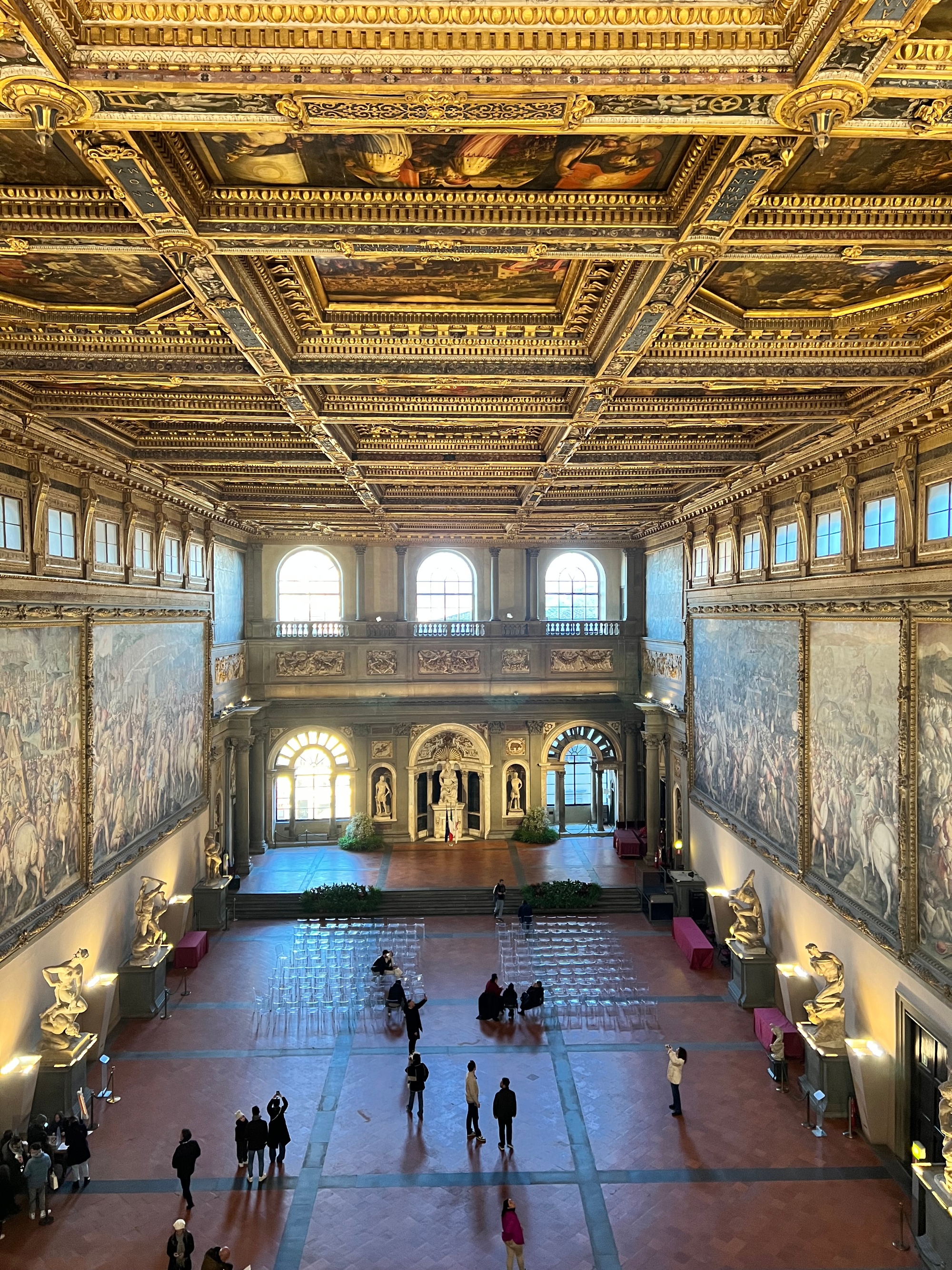 Salone dei Cinquecento in Palazzo Vecchio con soffitto dorato a cassettoni, grandi affreschi rinascimentali e visitatori nella sala.