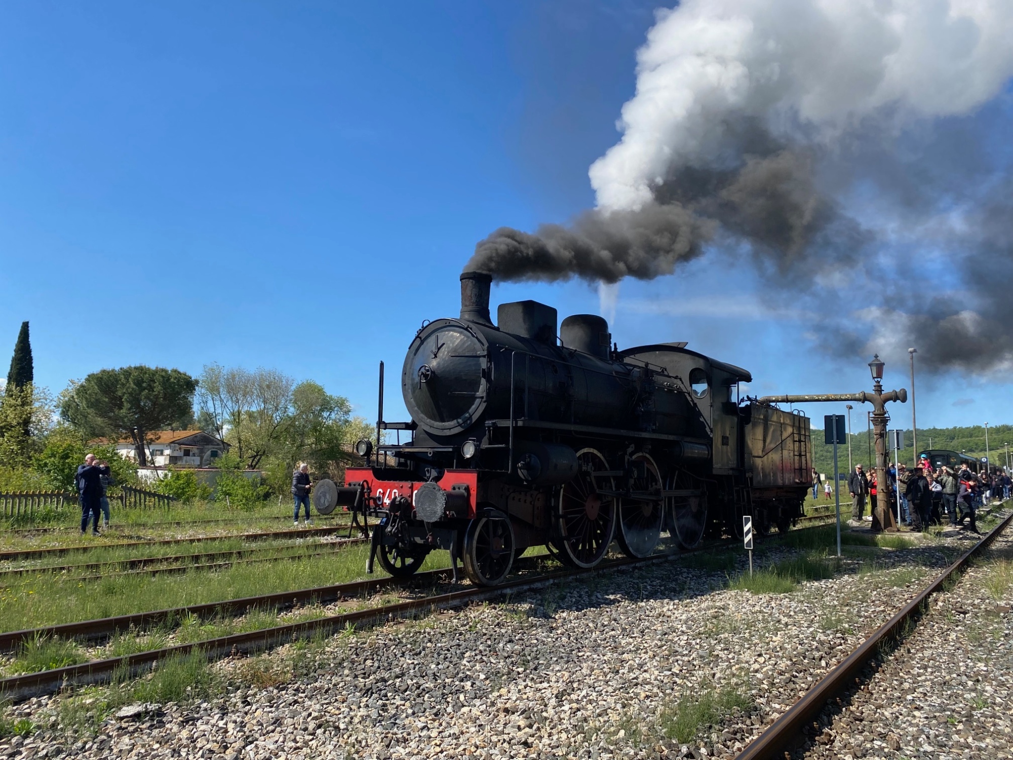Locomotiva a vapore nera con dettagli rossi ferma sui binari di una stazione toscana, con il fumo che si alza dal camino verso il cielo azzurro, mentre numerosi visitatori osservano la scena lungo i binari, nel contesto del Treno Natura che collega Siena a Trequanda attraverso le Crete Senesi e la Val d'Orcia