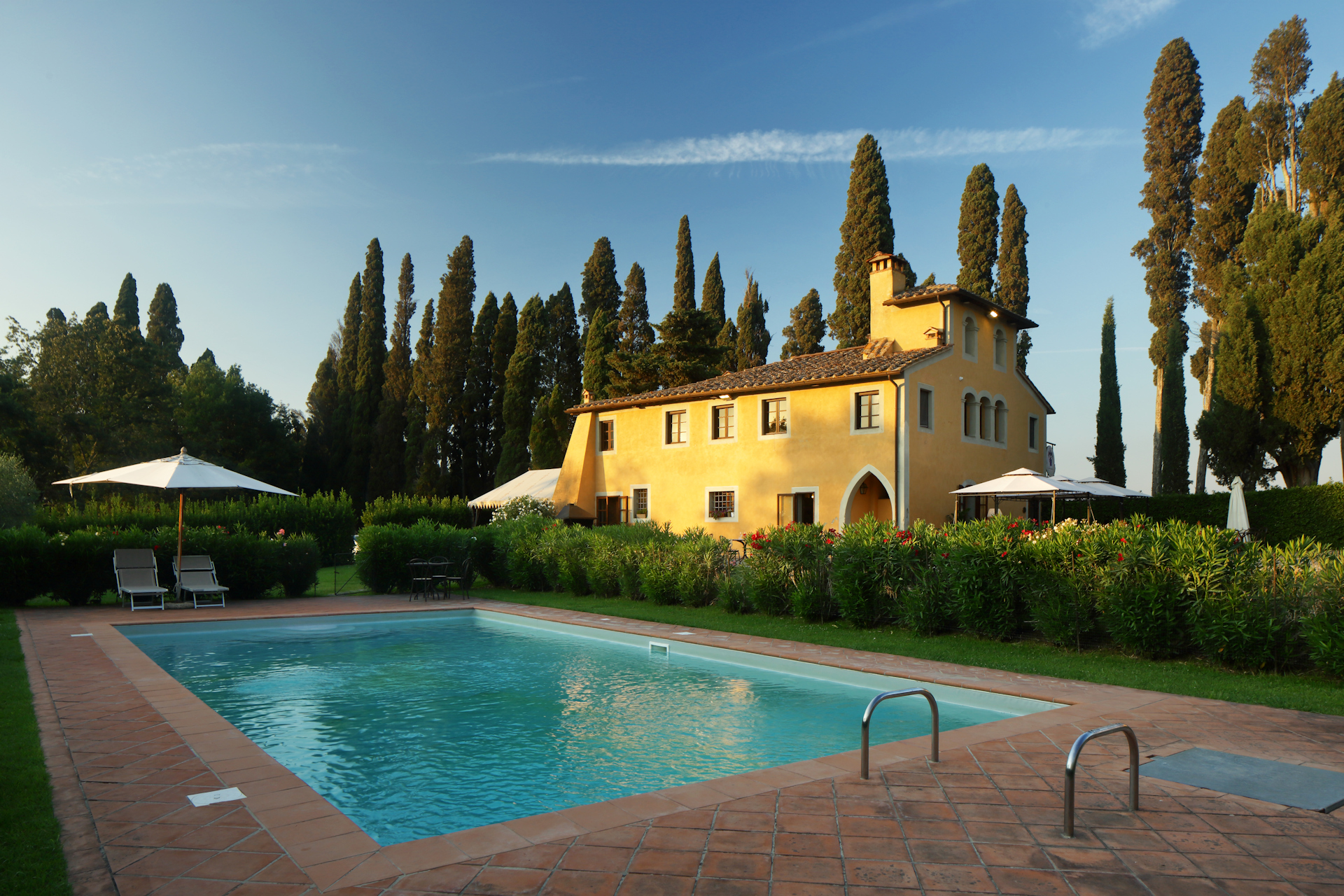 Agriturismo in Toscana con facciata gialla e piscina esterna, circondato da un giardino con solarium in cotto e un tipico viale di cipressi.