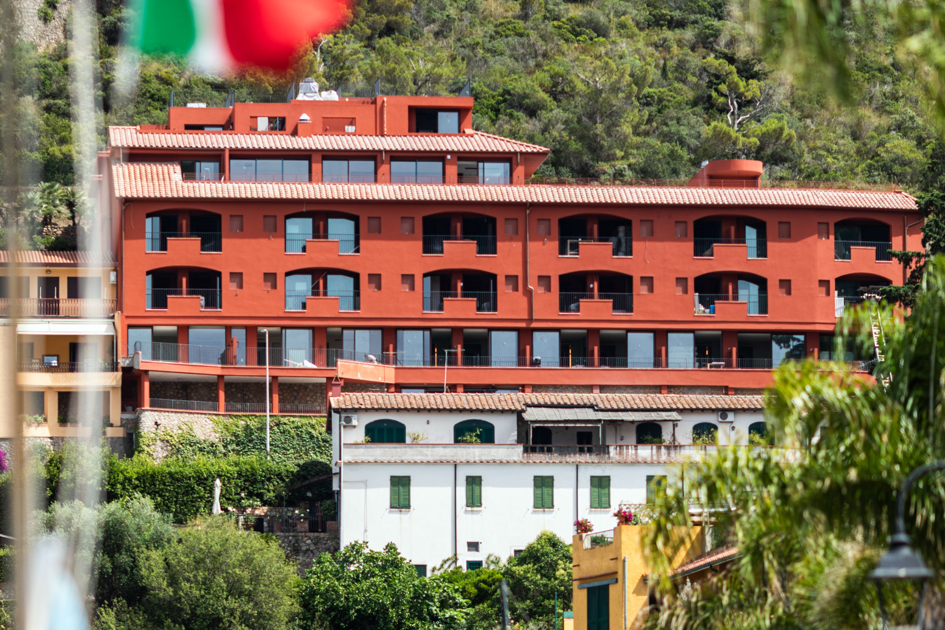 facciata esterna del boutique hotel LA ROQQA, situato a Porto Ercole. L'edificio principale si distingue nettamente per il suo vivace intonaco color rosso terracotta o rosso pompeiano, ed è strutturato su più livelli digradanti che seguono il profilo della collina retrostante, ricca di vegetazione spontanea e alberi. La facciata è ritmata da una serie ordinata di ampie logge e balconi ad arco scuro con ringhiere nere, appartenenti alle camere e alle suite, mentre l'ultimo piano ospita grandi vetrate e una terrazza panoramica. In basso, la struttura si inserisce nel contesto urbano del borgo marinaro, parzialmente coperta da alberi in primo piano e affiancata da edifici storici più bassi dalle tonalità bianche e gialle.