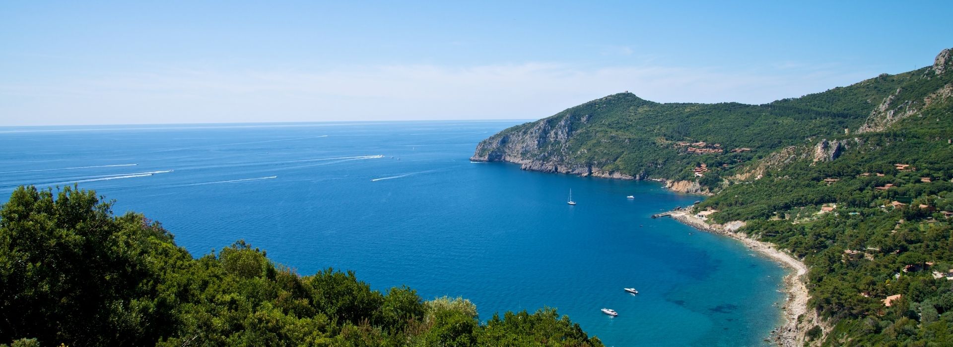 Panoramic view of the Monte Argentario promontory with its rocky coast covered in Mediterranean scrub, the blue sea with sailboats and motorboats, and the clear sky