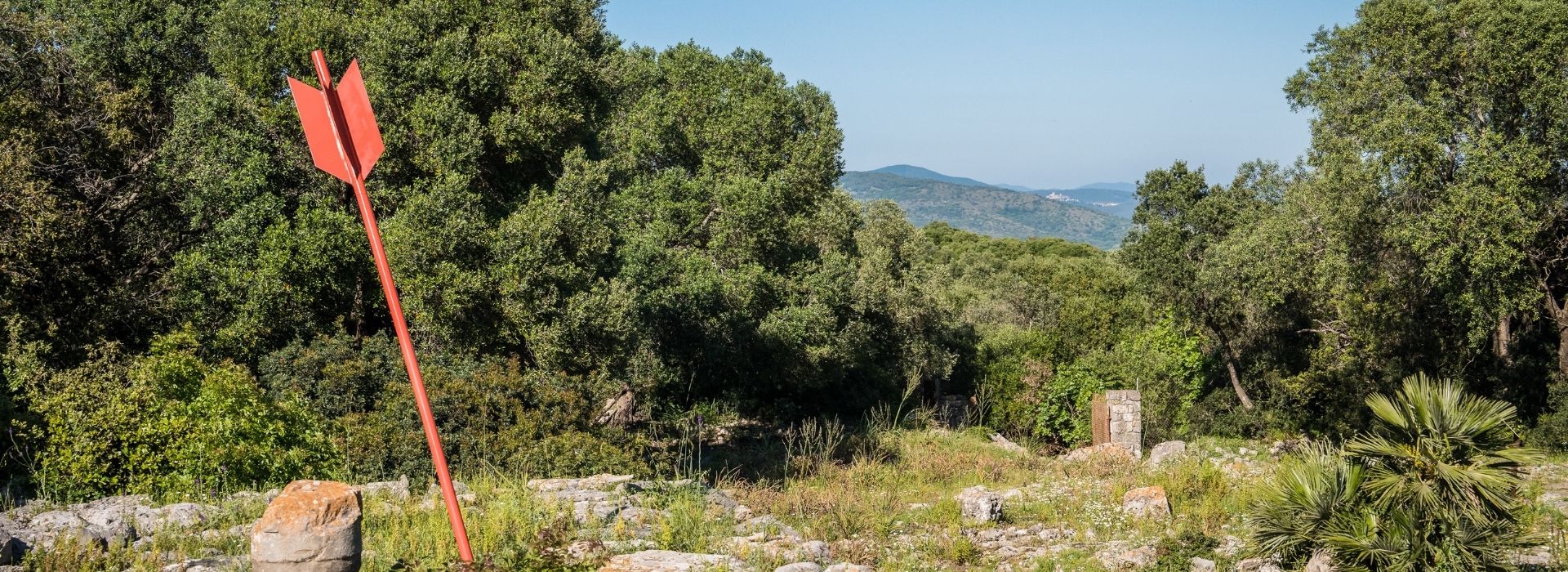 Segnaletica rossa a freccia per il sentiero e-bike immersa nella macchia mediterranea, con vista panoramica sulle colline dell'entroterra e un borgo in lontananza, tour in e-bike tra Argentario, Isola del Giglio e Laguna di Orbetello