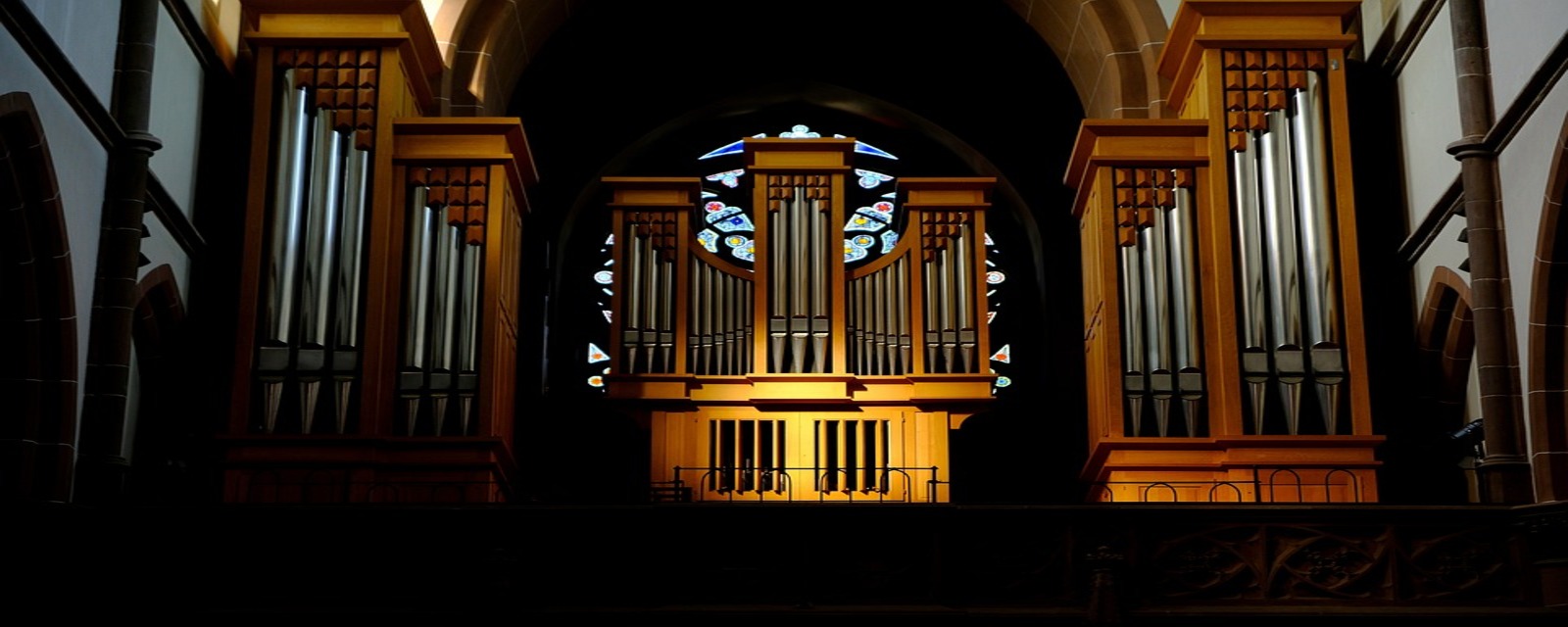 Organo a canne con struttura in legno illuminata, davanti a un rosone con vetrate colorate all’interno di una chiesa.