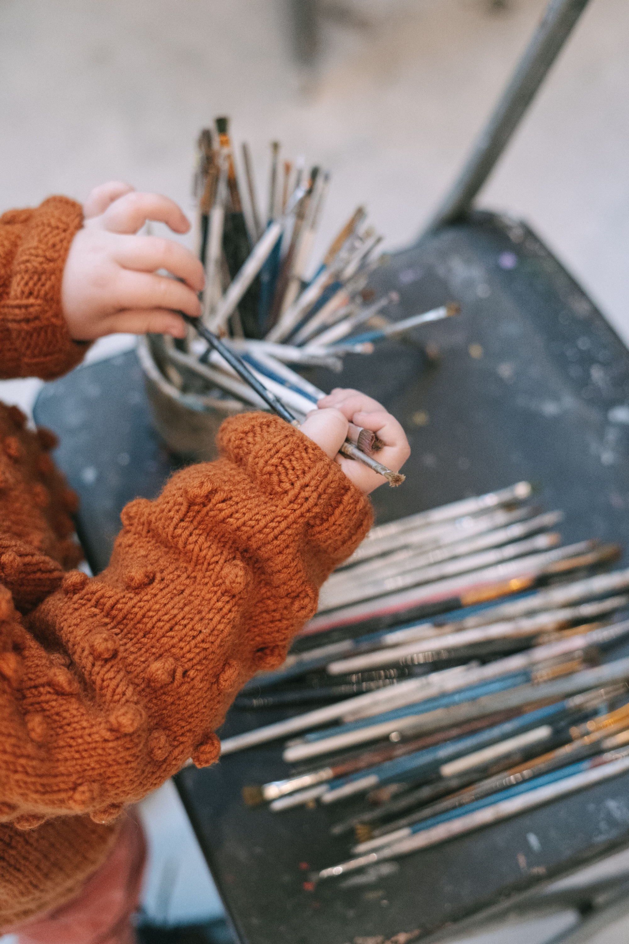 Mani di una bambina con un maglione arancione che scelgono alcuni pennelli tra quelli disposti su un tavolo da lavoro.