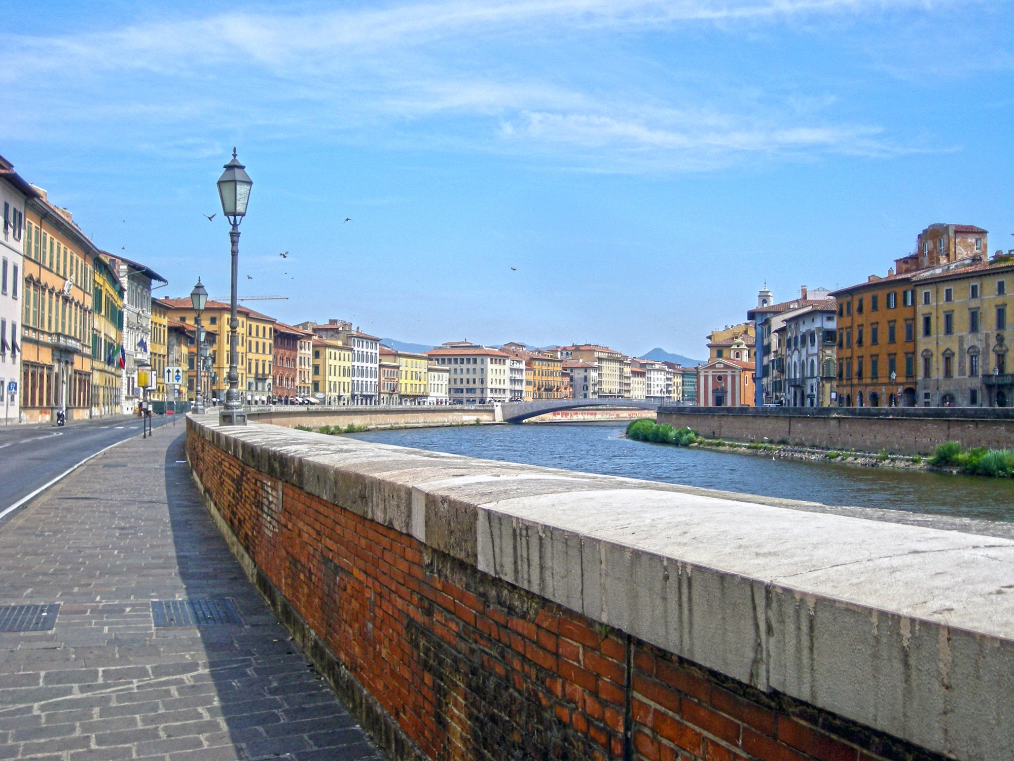 Los Lungarni de Pisa: en primer plano, el muro de ribera de ladrillo y el paseo enlosado; en el centro, el río Arno, enmarcado por históricos edificios de colores, y un puente al fondo.