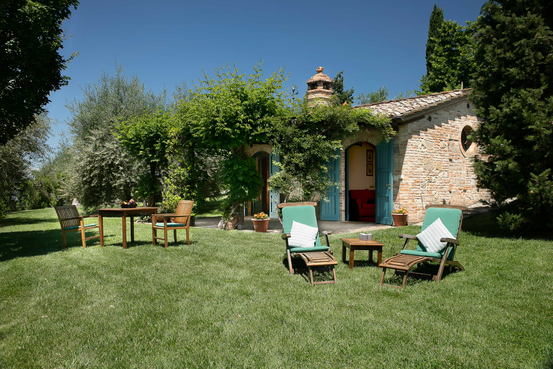 The entrance of an apartment with a garden in front where there are chairs, deckchairs and wooden tables