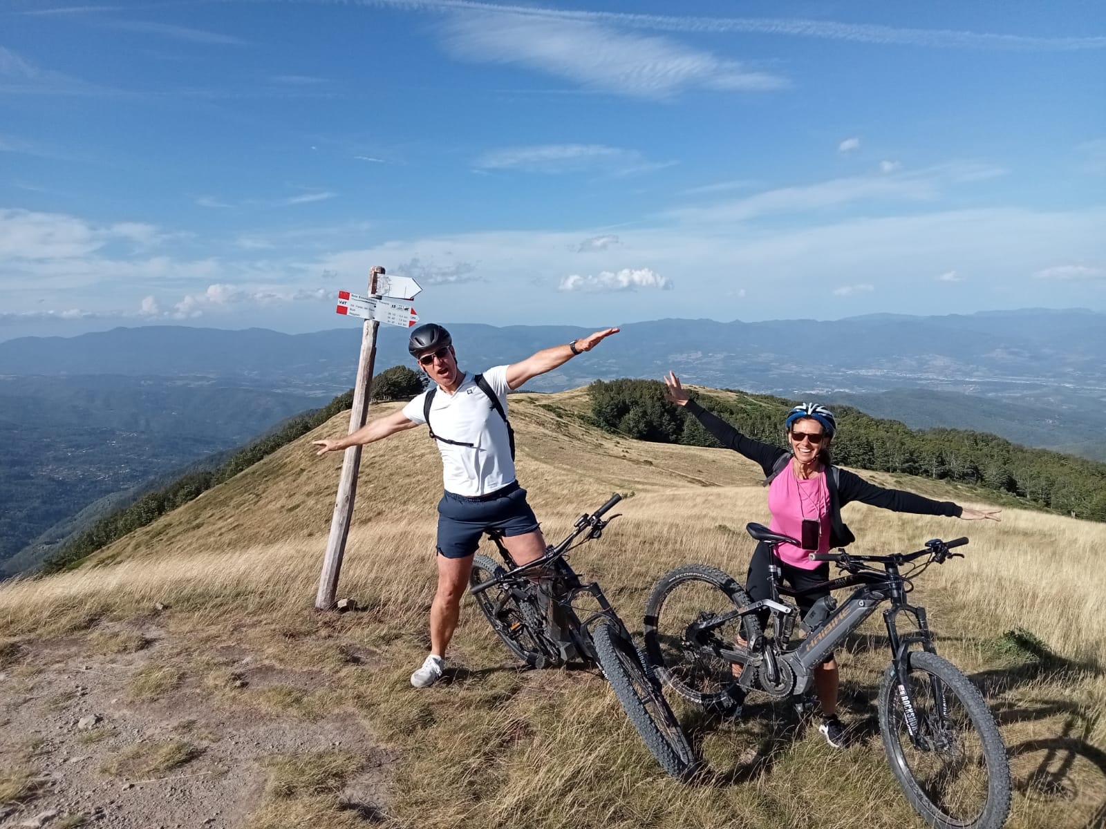 Due ciclisti con caschi e e-bike in posa con le braccia aperte sulla cima erbosa del Pratomagno, con vista panoramica a 360° sul Casentino e il Valdarno sullo sfondo