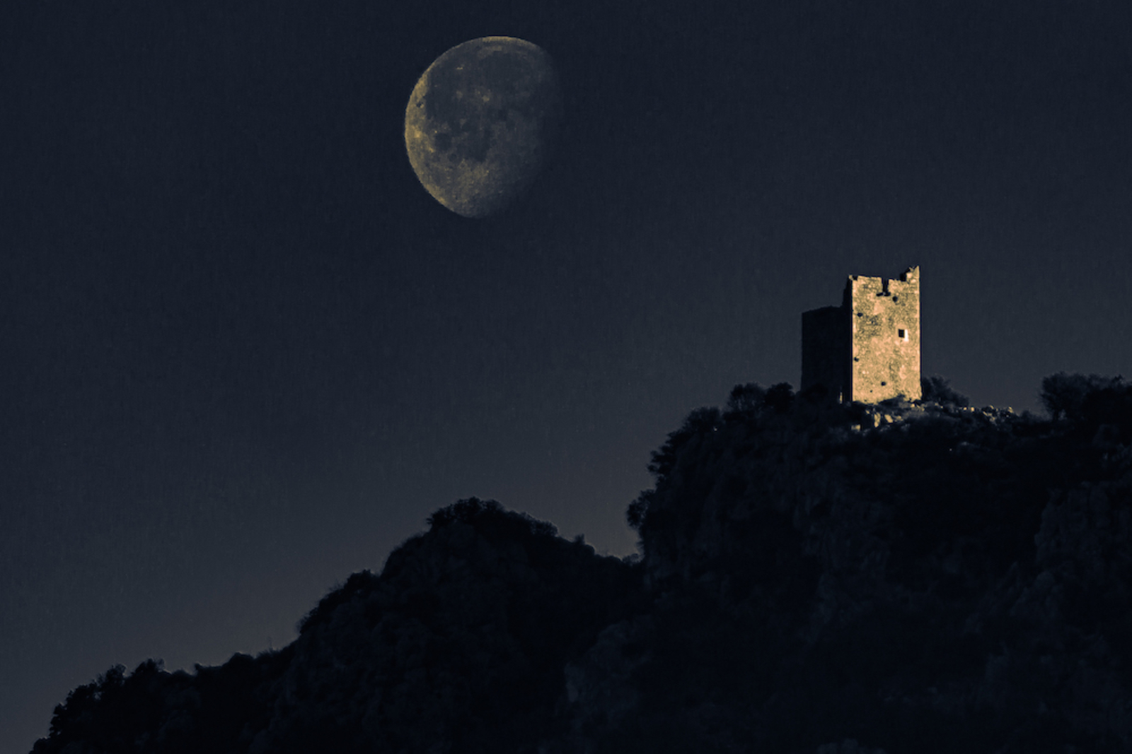 Grande luna gibbosa sospesa nel cielo notturno blu scuro sopra una collina rocciosa con un'antica torre di avvistamento in pietra illuminata dalla luce lunare e la sagoma scura della macchia mediterranea in primo piano