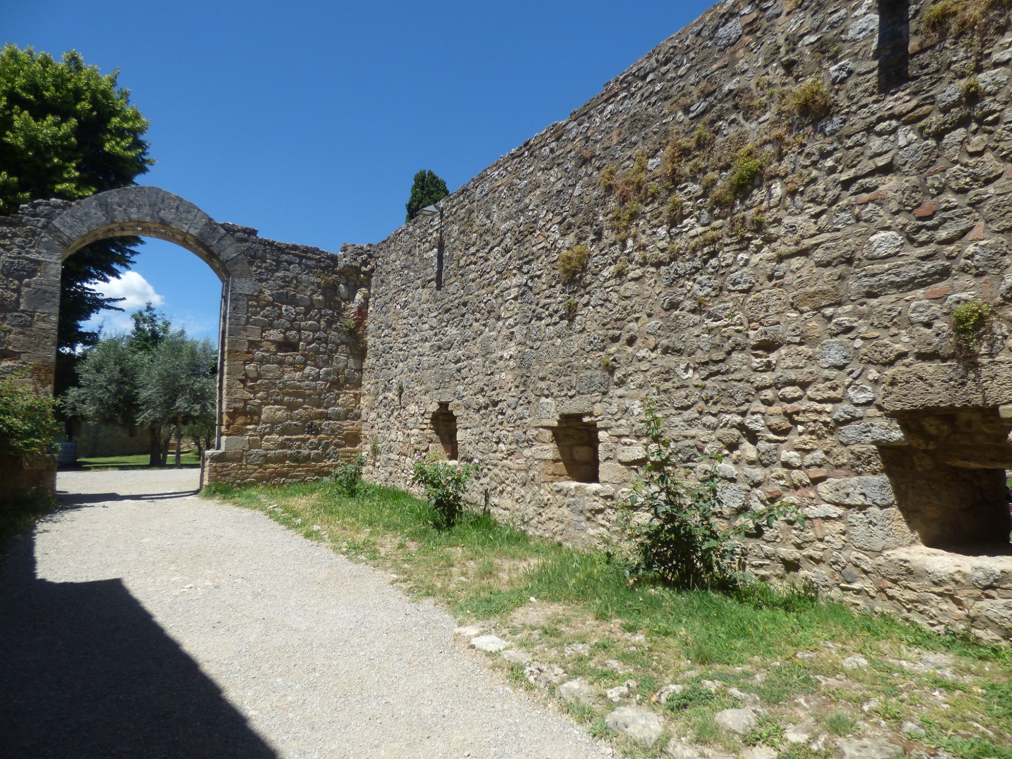 Un détail des anciennes structures défensives, avec une longue enceinte en pierre dotée de meurtrières et d’un grand portail en arc donnant sur un parc intérieur ombragé.
