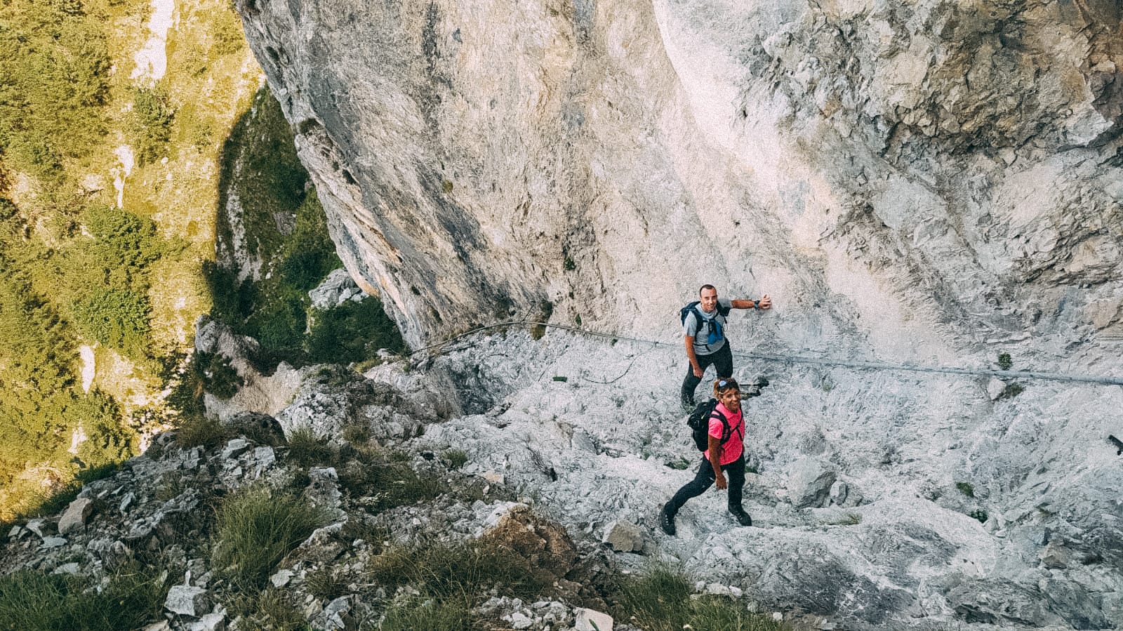 Due escursionisti con zaino percorrono un sentiero roccioso e ripido lungo una parete di marmo delle Alpi Apuane, agevolati da cavi metallici di sicurezza, con vegetazione verde visibile sul versante sottostante