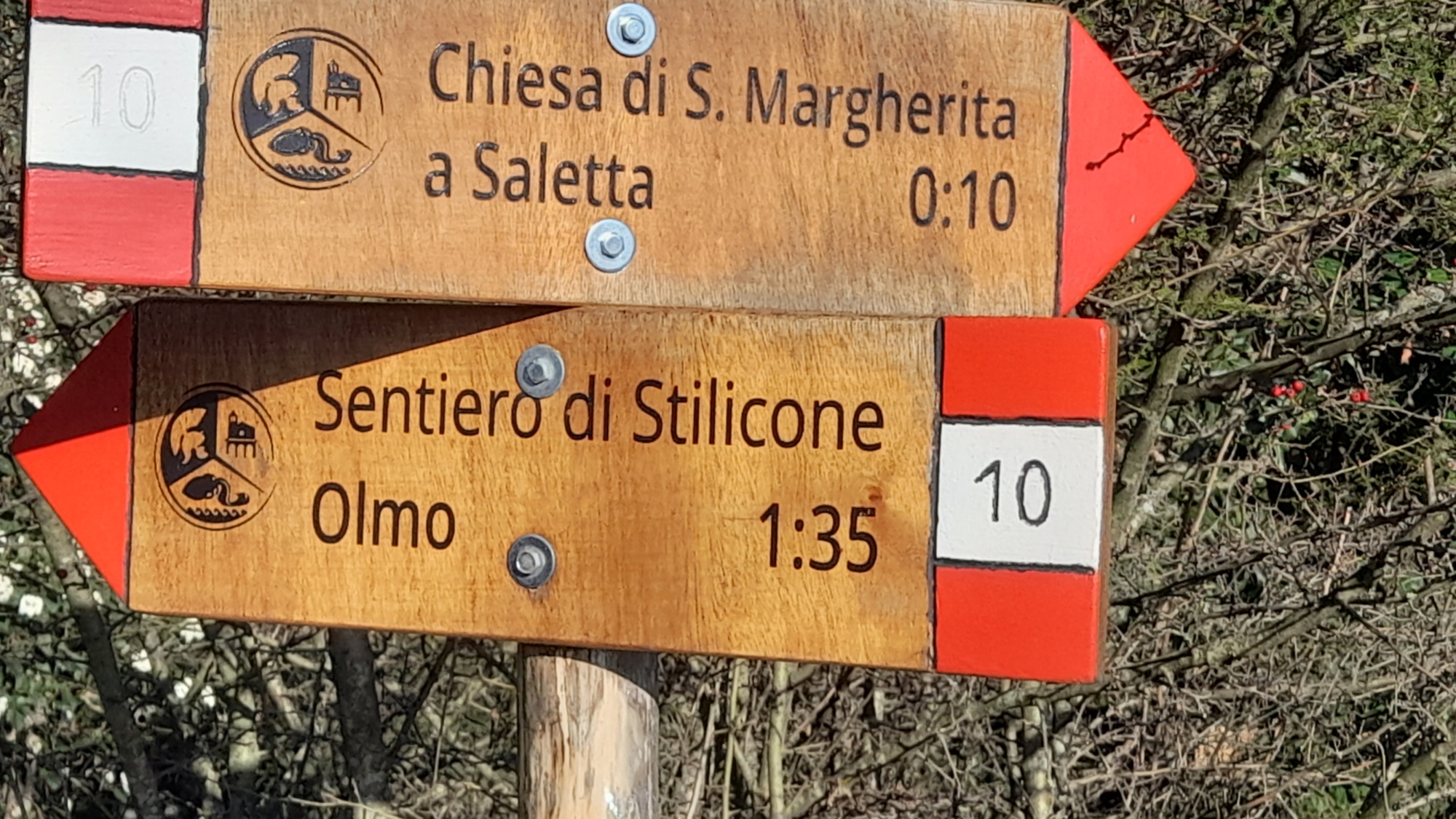 Wanderschild aus Holz mit roten Pfeilen, die auf zwei Routen verweisen: rechts auf die Kirche Santa Margherita in Saletta und links auf den Stilicho-Weg Richtung Olmo