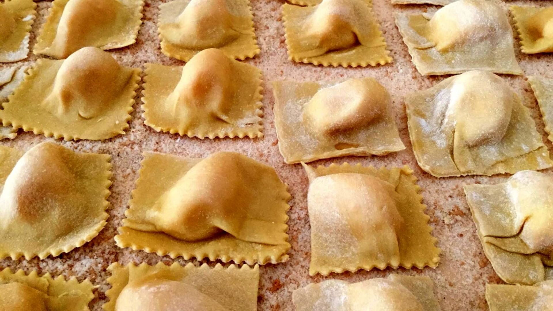Una fila ordinata di tortelli (o ravioli) di pasta fresca fatti a mano, disposti su una superficie leggermente infarinata. I bordi della pasta sono seghettati e il ripieno al centro è ben visibile e rigonfio.