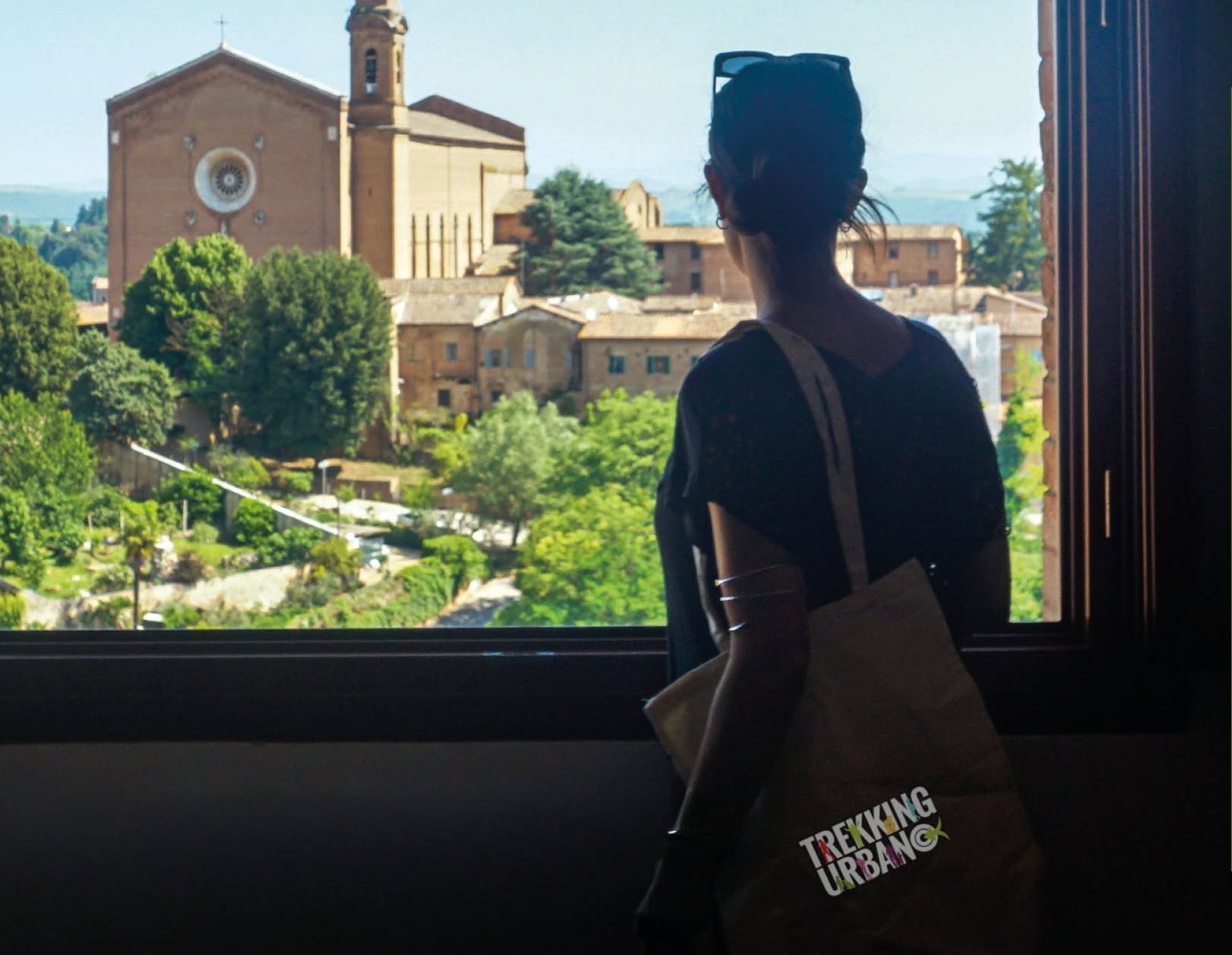 Persona di spalle osserva il centro storico di Siena da una finestra, con una chiesa e il paesaggio urbano sullo sfondo.