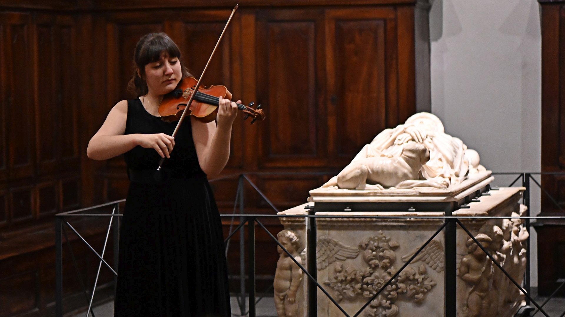 Una musicista con abito scuro suona il violino davatnìanti al monumento funebre di Ilaria del Carretto