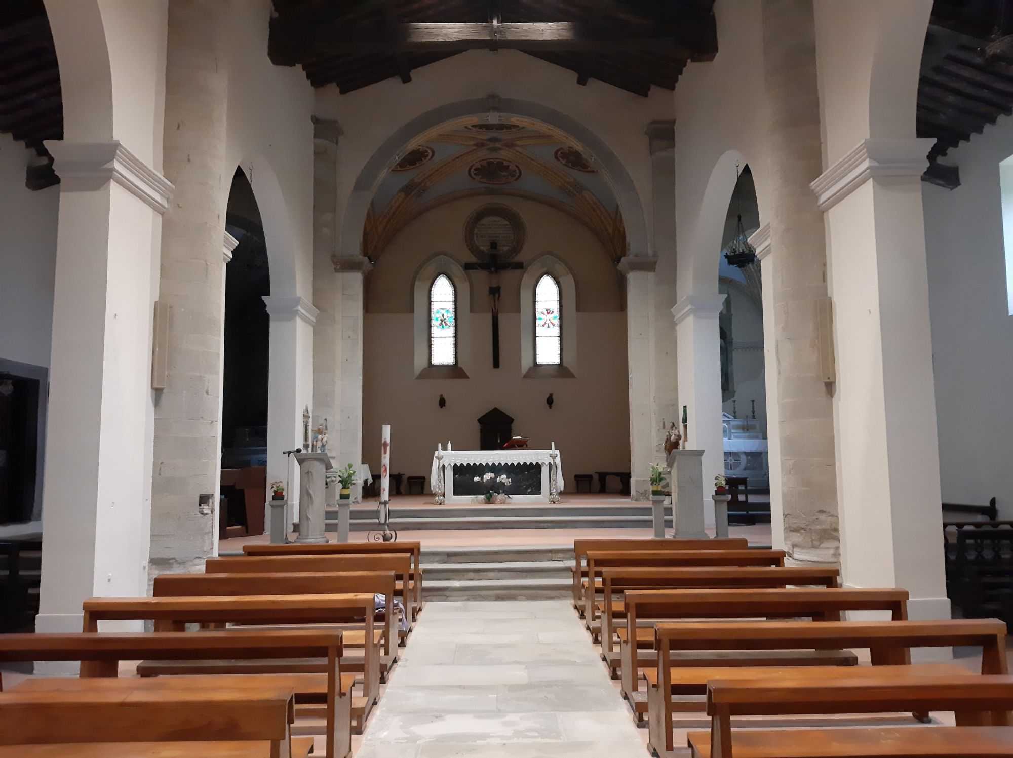 Interno della chiesa che mostra la navata centrale con file di panche in legno, grandi colonne bianche che sorreggono archi, un soffitto a capriate lignee e l'altare maggiore sullo sfondo sormontato da un crocifisso e due finestre con vetrate decorate