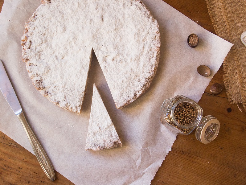 Panforte coated with powdered sugar, from which a slice is missing. Also glimpsed on the table are the spices used in the recipe.